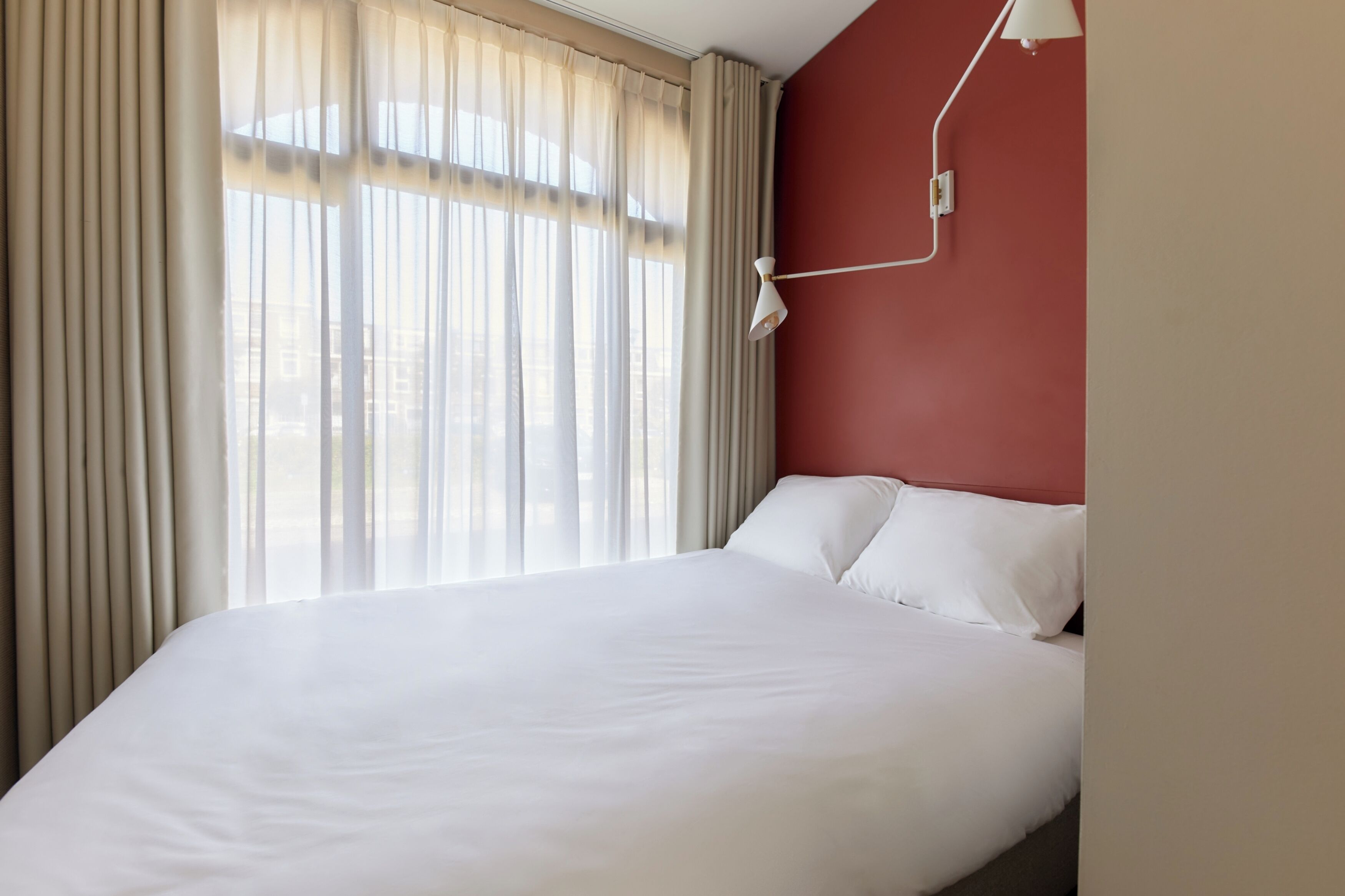 A hotel room with a large bed, beige curtains, a white lamp, and a red wall.