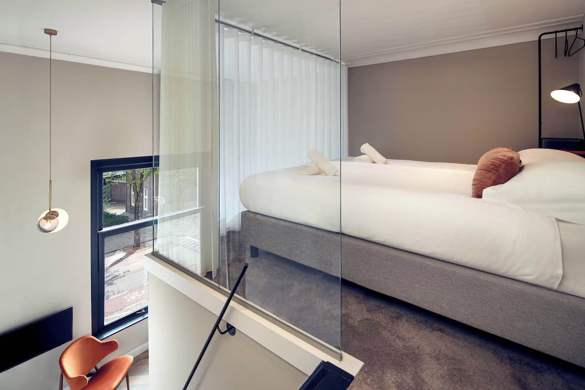 A modern bedroom with a large bed, a glass partition, a chair, and a view of the street outside.