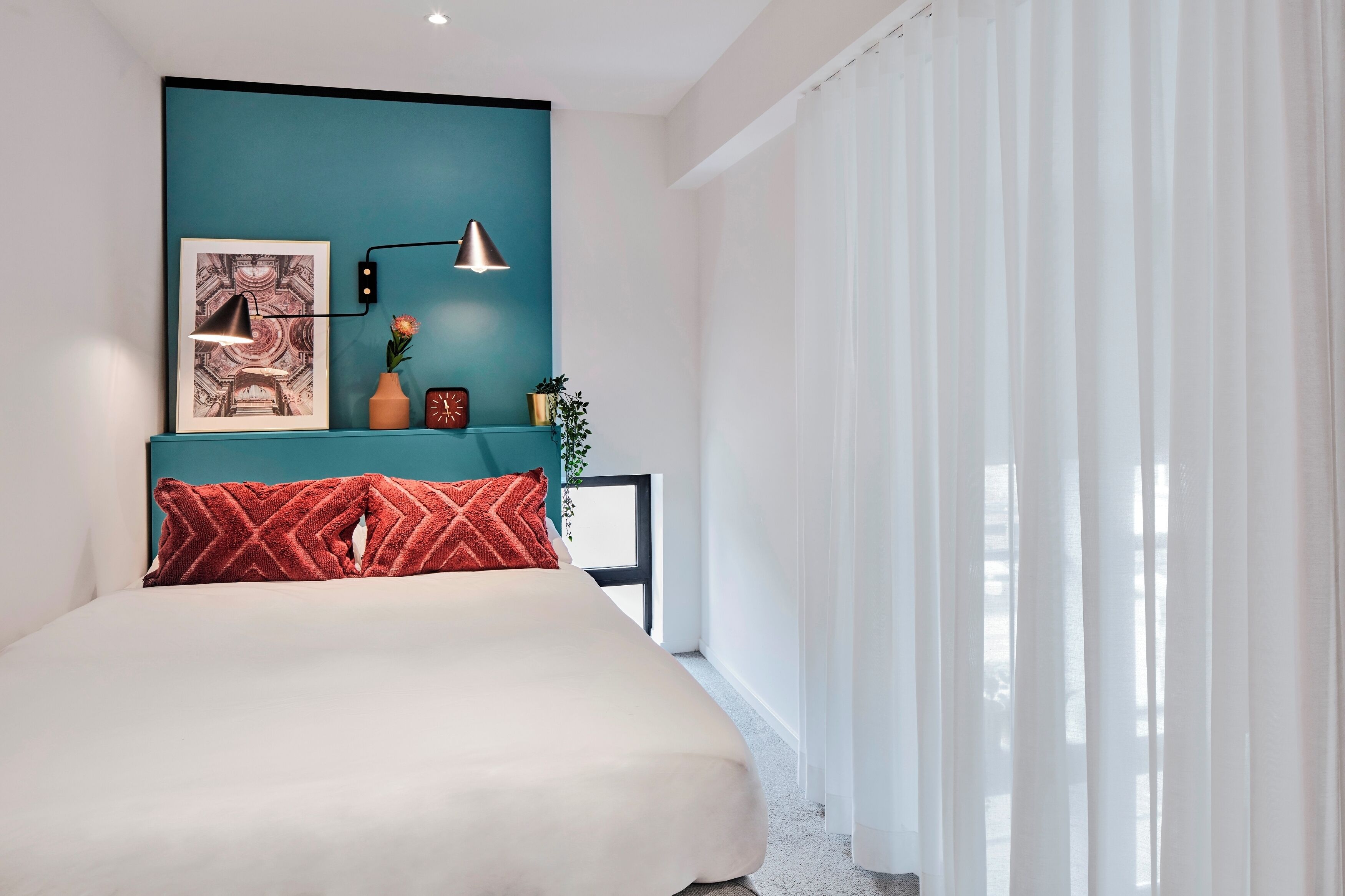 A bedroom with a teal accent wall, a bed with a red patterned pillow, a framed picture, a small plant, and a wall-mounted lamp.