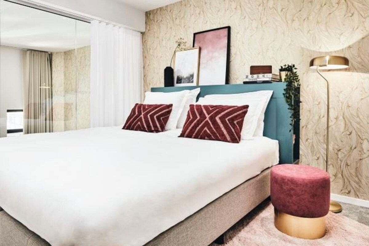 Un dormitorio moderno con una cama grande, dos almohadones decorativos, una mesita de noche, una lámpara y una pared con textura.