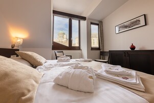 Quarto de hotel com cama, toalhas, luminária, janela com vista para um edifício e um quadro na parede.