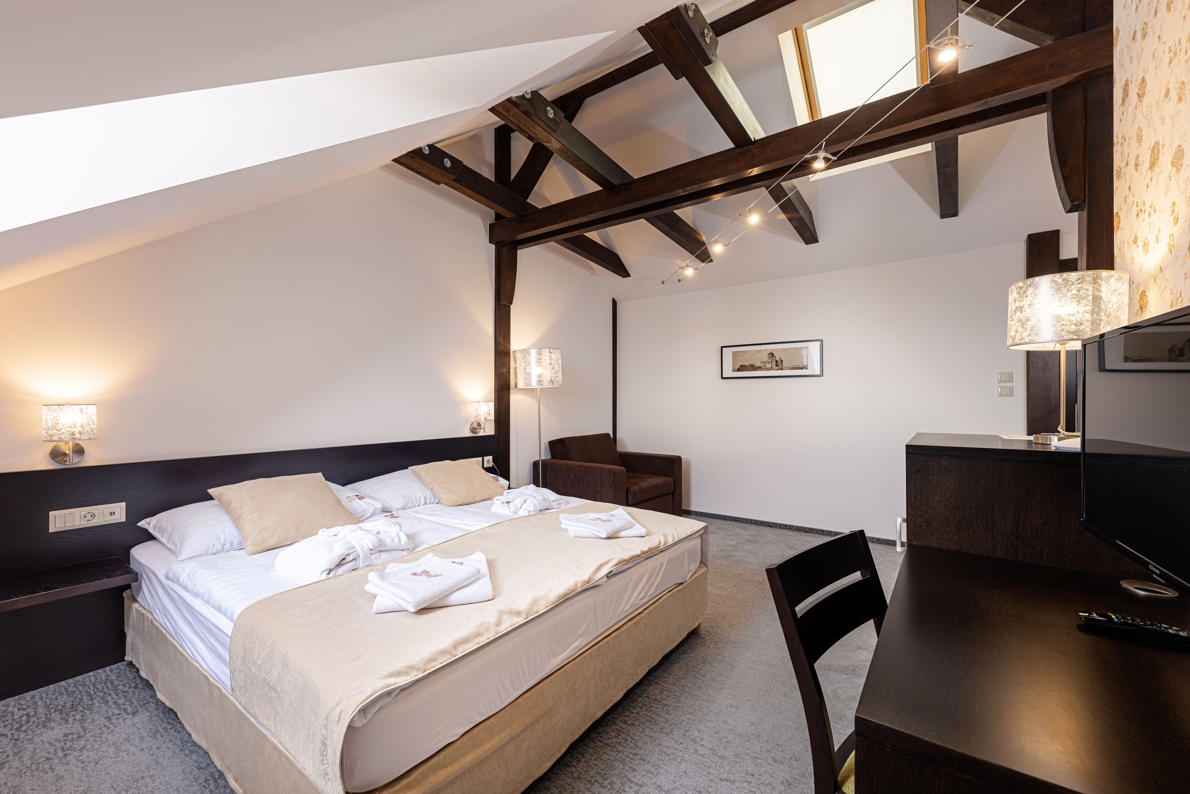 Habitación de hotel moderna con una cama grande, un escritorio y una silla. El techo presenta vigas a la vista y una lámpara sobre el escritorio.