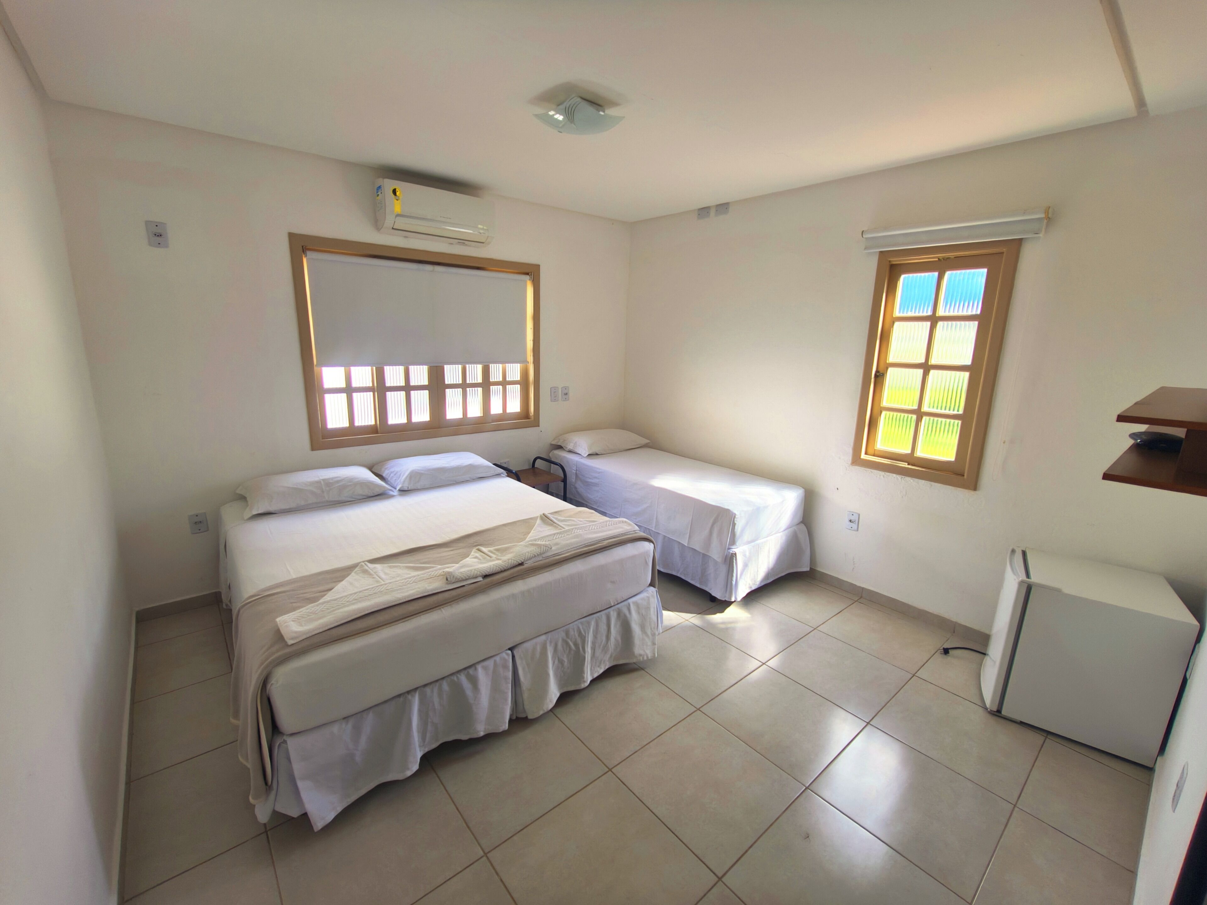 Una habitación con dos camas, un escritorio pequeño, un refrigerador y una ventana con persianas.