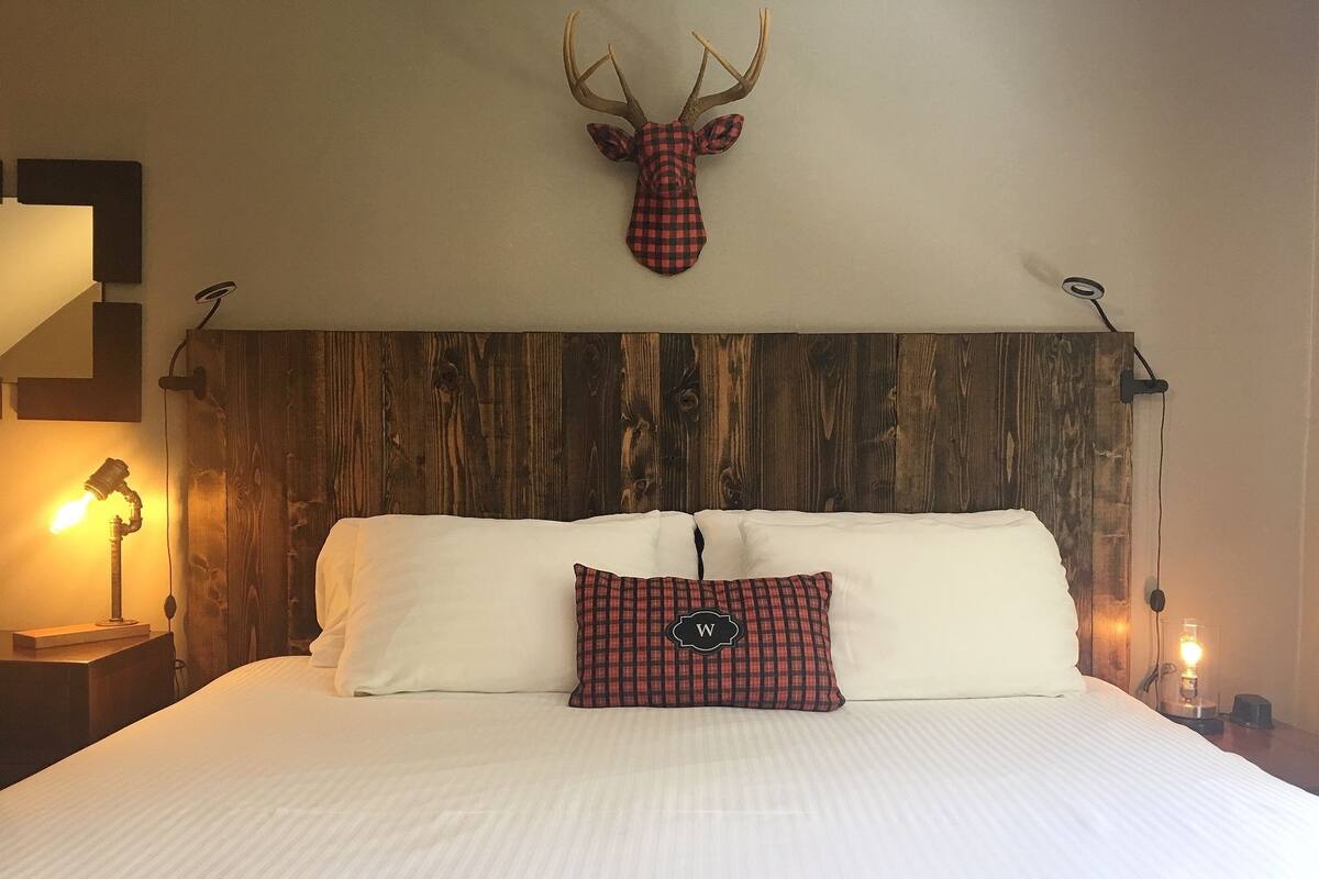 A bedroom with a wooden headboard, white bedding, a checkered pillow, and a mounted deer head.