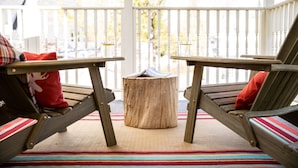Deux chaises en bois avec des coussins rouges, placées sur une véranda où se trouvent un tapis à rayures, une petite table en bois, et où l’on peut admirer, au loin, des arbres et une clôture blanche.