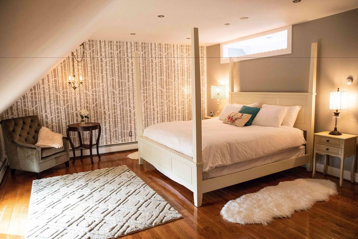 A bedroom with a four-poster bed, a sitting area with a chair and table, and a large window providing natural light.