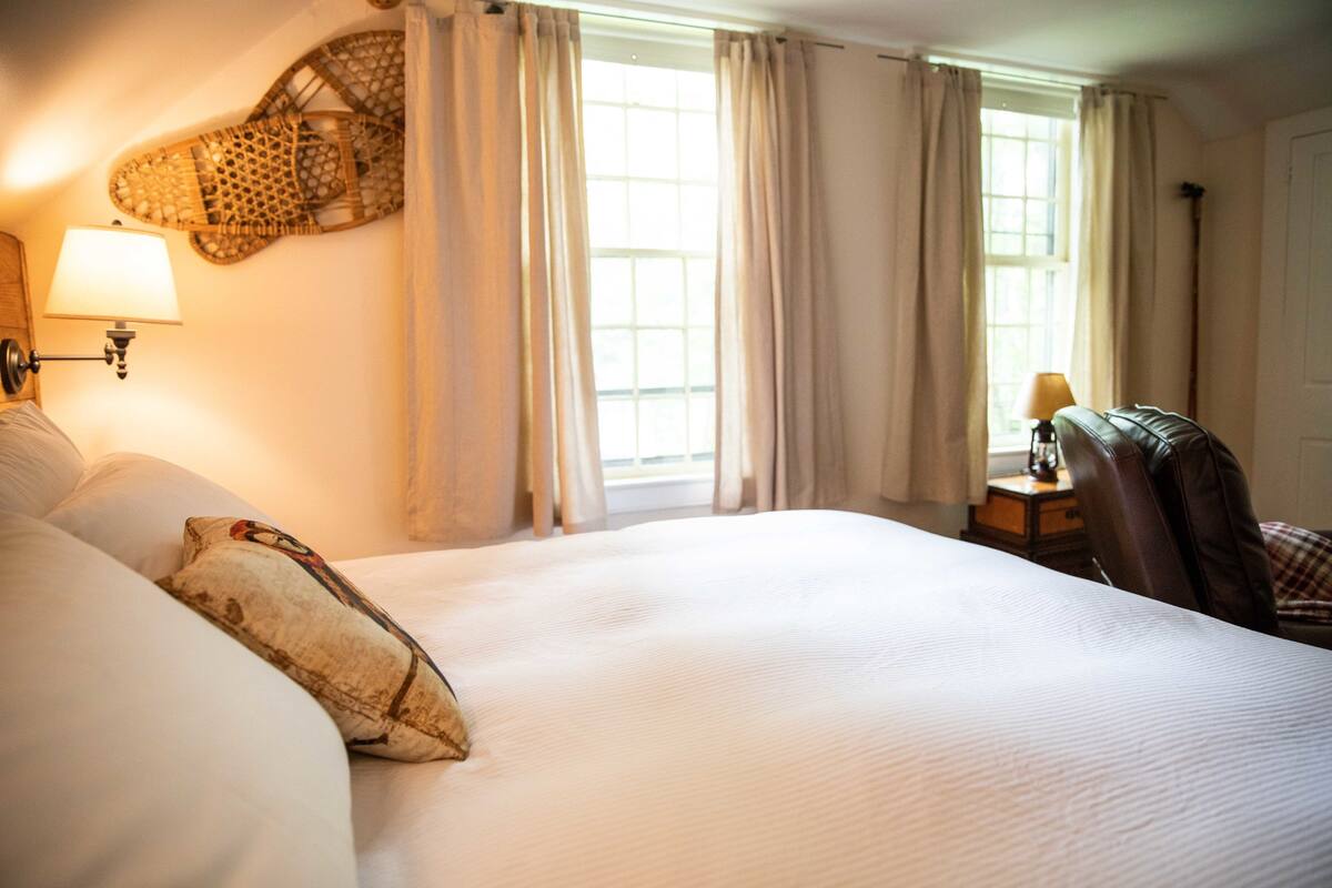 A hotel room with a large bed, two bedside lamps, a chair, a small table, and a window with curtains.