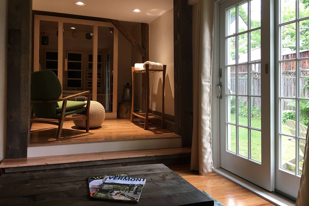 A cozy room with a wooden floor, a green chair, a wooden table with a magazine, and a large window with a view of a backyard.