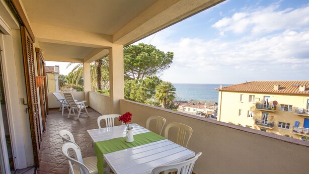 Un balcone con tavolo e sedie, vista su una zona costiera con edifici e alberi.