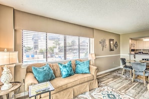 A living room with a beige sofa, teal cushions, a small table with a lamp, and a view of the outside through large windows.