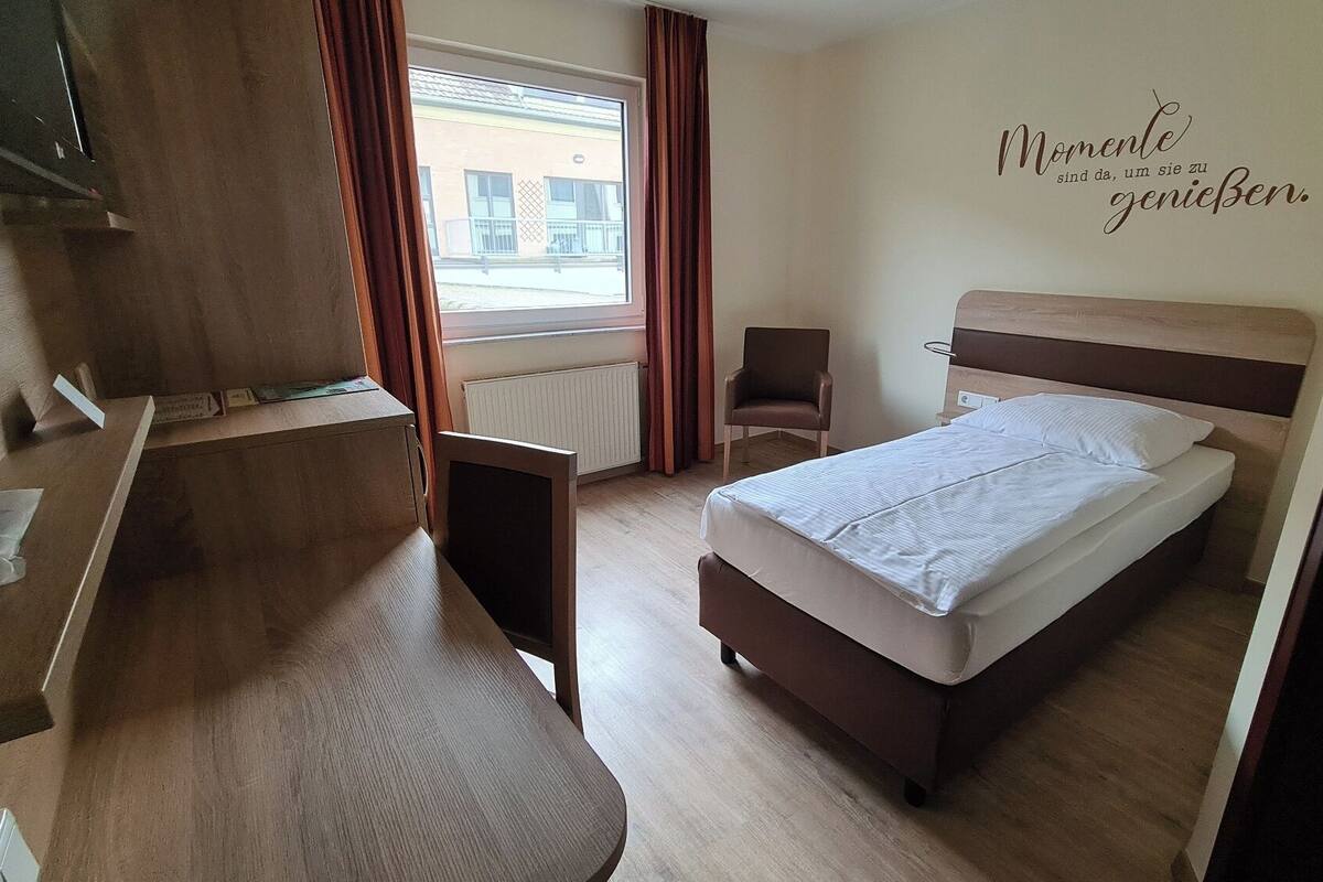 Quarto de hotel com cama, escrivaninha, cadeira e janela com cortinas.