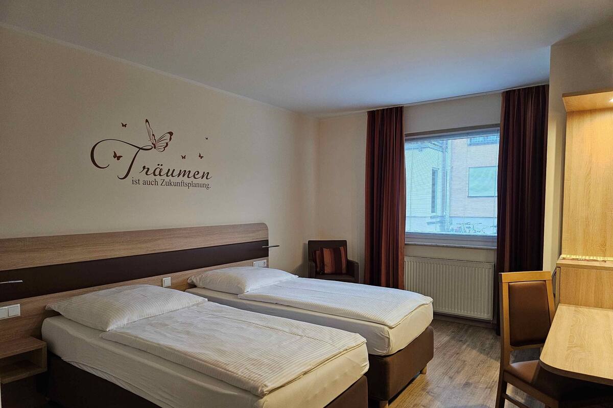 Quarto de hotel com duas camas, uma escrivaninha, uma cadeira, uma janela com cortinas e um adorno na parede.