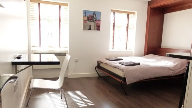 A small, well-lit room with a bed, a desk, and a chair. There are two windows providing natural light. - SS Property Hub - City of London Studio Apartment 12 (London)