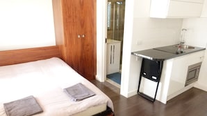 A bedroom with a bed, a wooden wardrobe, a small kitchenette with a sink and microwave, and a shower area. - SS Property Hub - City of London Studio Apartment 12 (London)