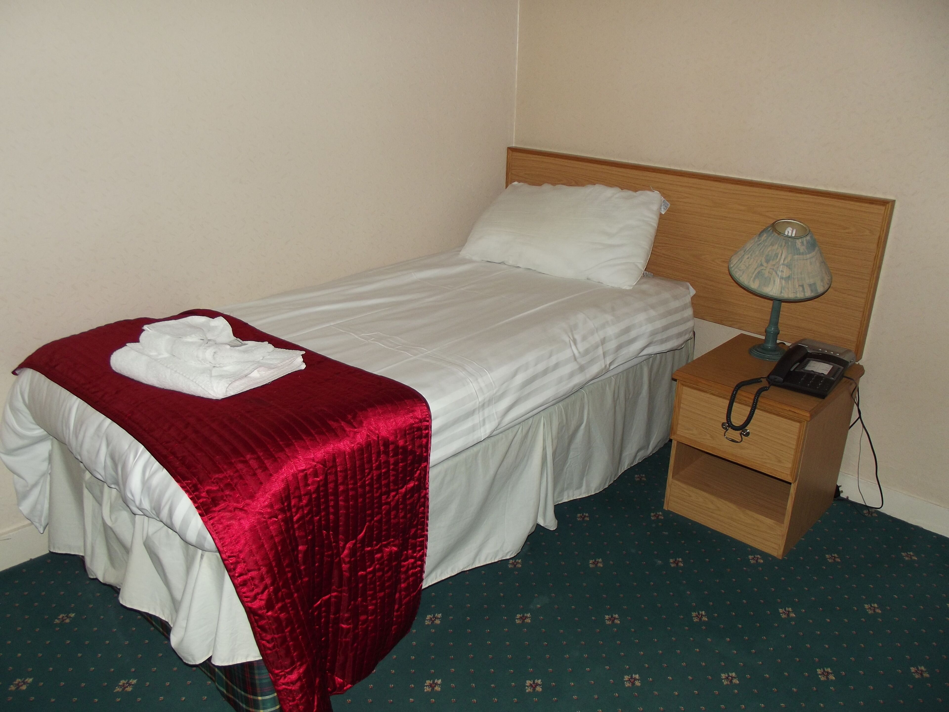 A neatly made bed with a red blanket and white pillows, a bedside lamp, and a phone on a wooden nightstand.