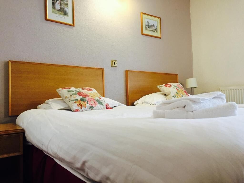 A hotel room with two beds, each with floral pillows and white linens. There are framed pictures on the wall and a lamp on the nightstand.