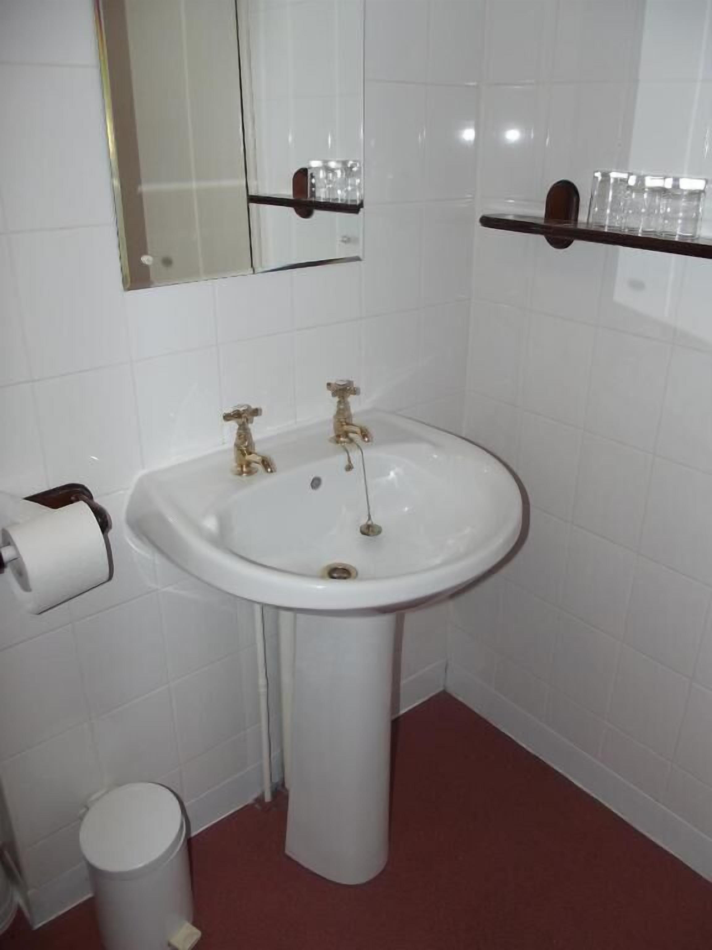 Une salle de bain comprenant un lavabo sur colonne, des toilettes, une poubelle, un miroir et une étagère avec des articles de toilette.