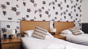 Hotellihuone, jossa on kaksi sänkyä, puiset päätylevyt, yöpöydät lampuineen ja kukallinen tapetti.