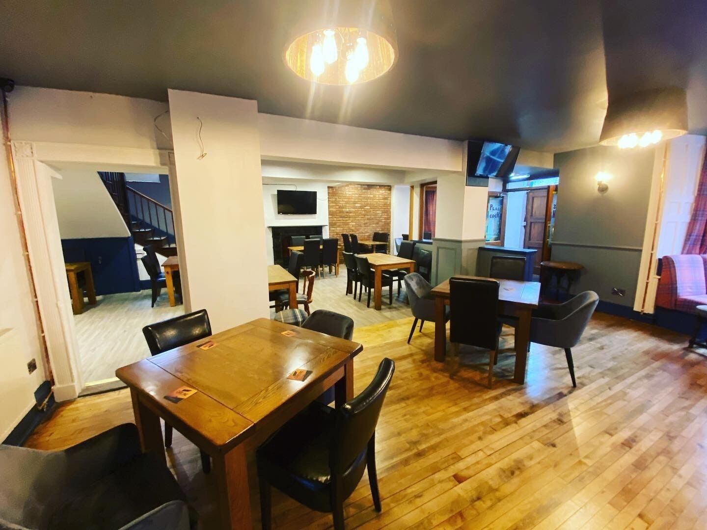 A cozy restaurant with wooden flooring, multiple tables and chairs, a TV mounted on the wall, and a staircase leading to another area.