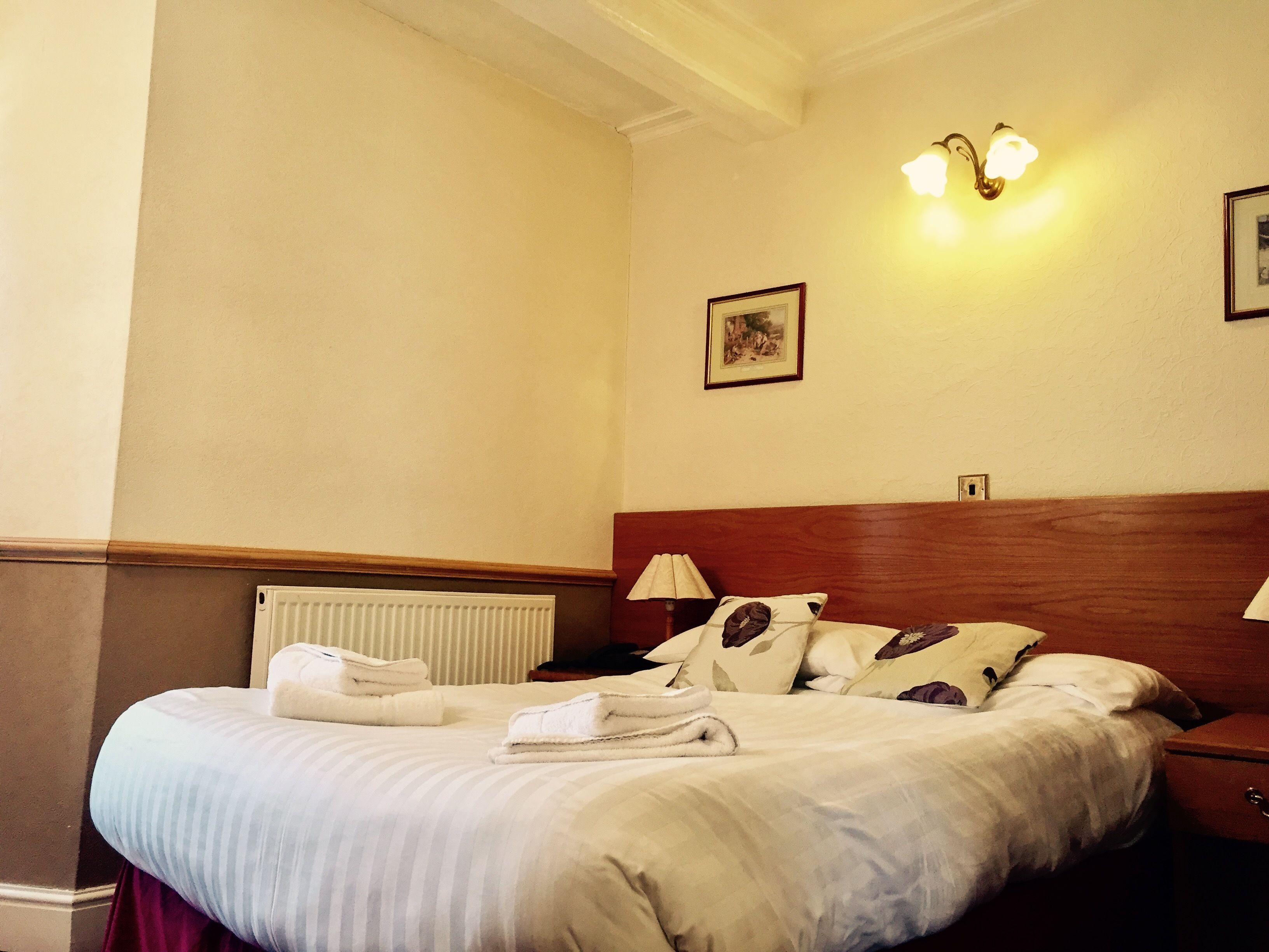 A hotel room with a neatly made bed, a radiator, a wooden headboard, and two wall-mounted lamps.