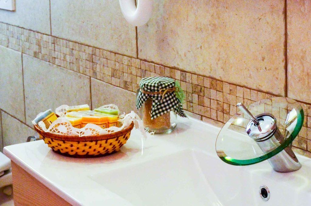 Un lavabo de salle de bain avec un porte-savon en verre, un panier en osier contenant des articles de toilette et un bocal en verre recouvert d’un tissu à carreaux.