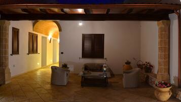 Eine Außenpatio-Terrasse mit Steinboden, Korbmöbeln, einem Tisch mit Lampe und einer großen Vase mit Blumen.