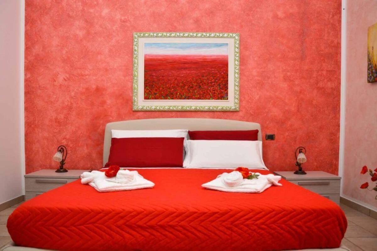 Una habitación de hotel con una colcha roja, dos almohadas, un cuadro enmarcado en la pared y una mesita con una lámpara.