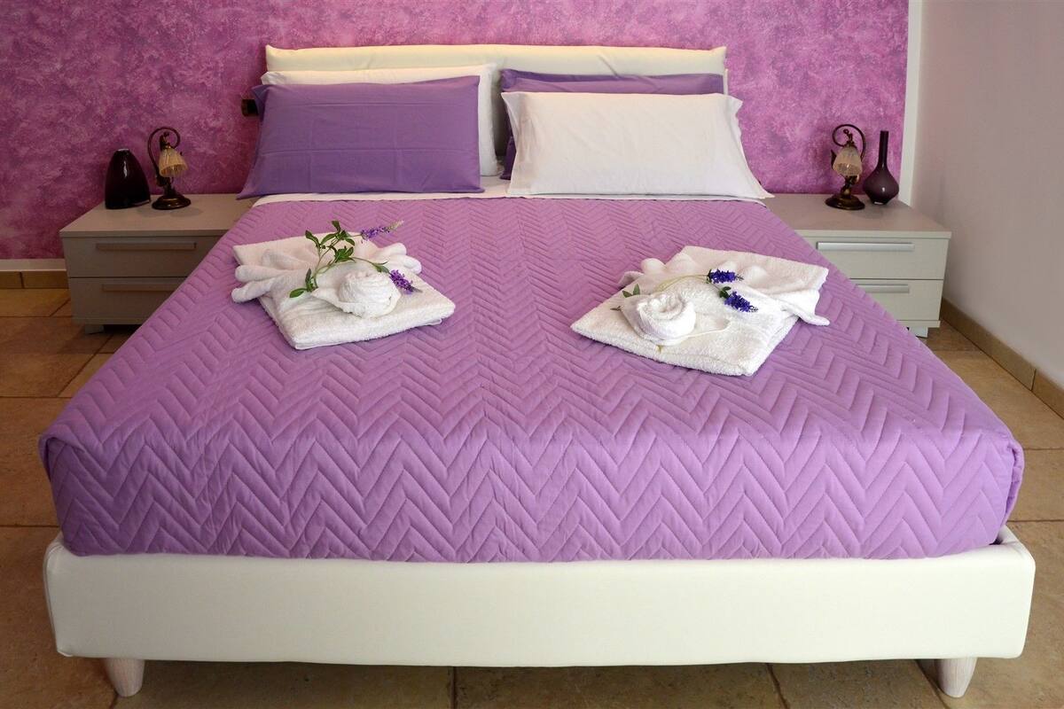 Una cama bien hecha con un edredón morado, dos toallas blancas con motivos de lavanda y almohadas a juego sobre una mesita de noche.