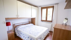 Ein Schlafzimmer mit Bett, Nachttisch, Kommode, einem Fenster mit Vorhängen und einer Heizung.