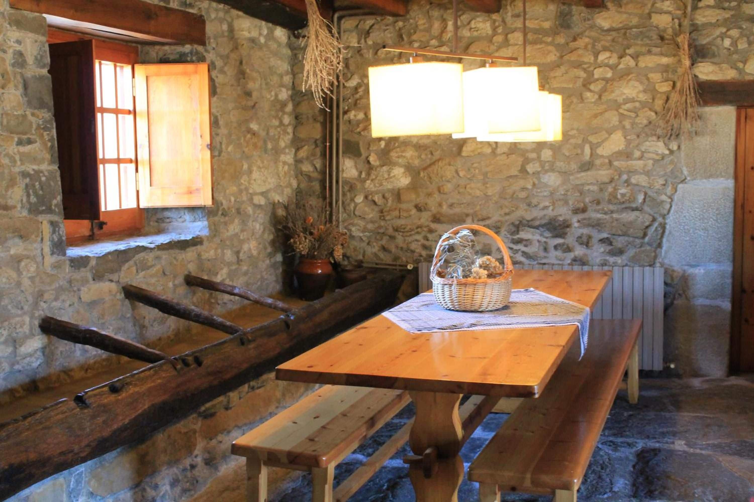 Interior rústico de pedra com mesa e bancos de madeira, janela com persianas e luminária pendente.
