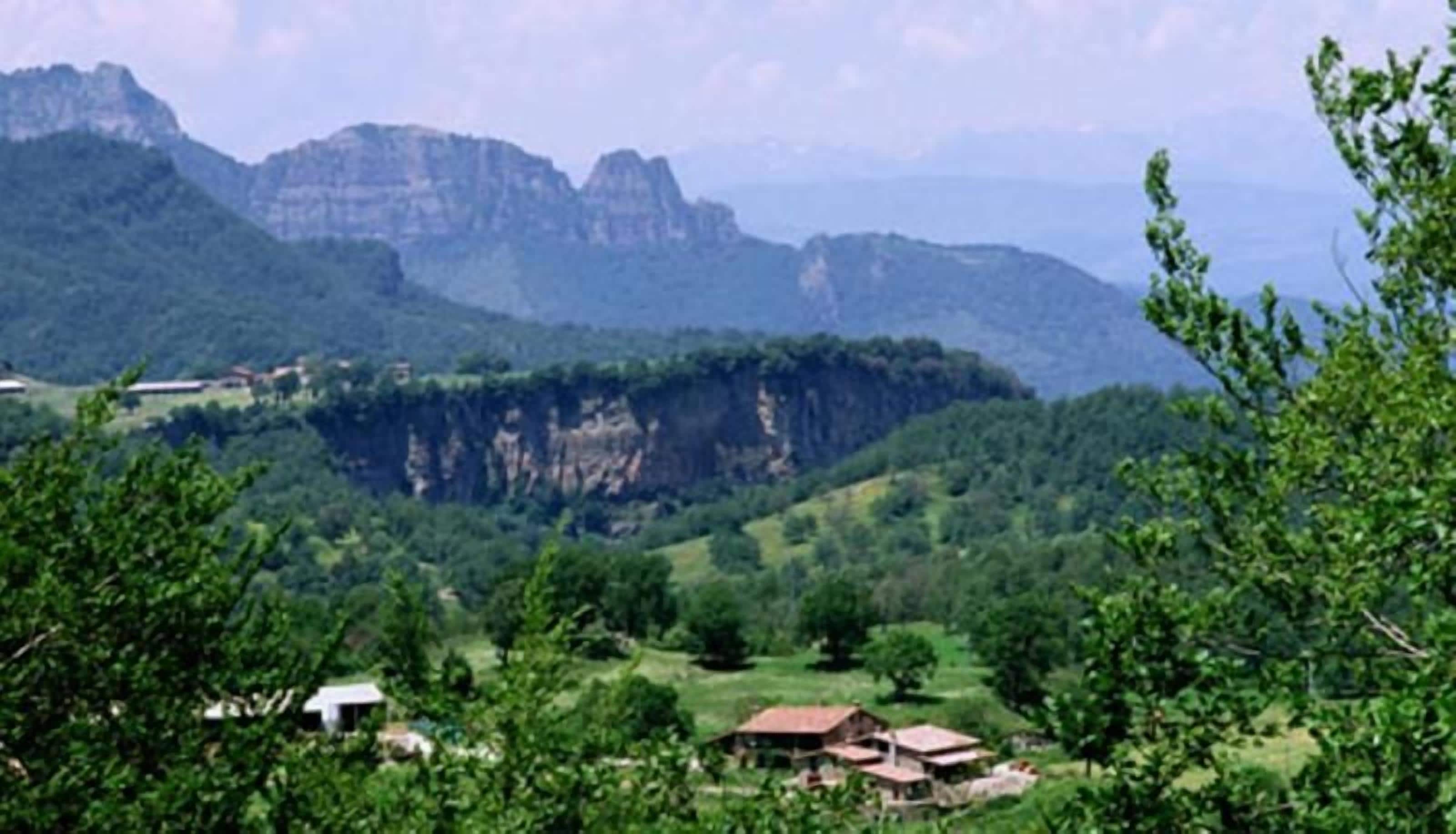 Paisaje rural con una casa rodeada de vegetación y montañas al fondo.
