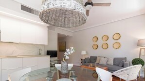 Un salon moderne avec une table à manger en verre, des chaises blanches, un canapé gris et un ventilateur de plafond.