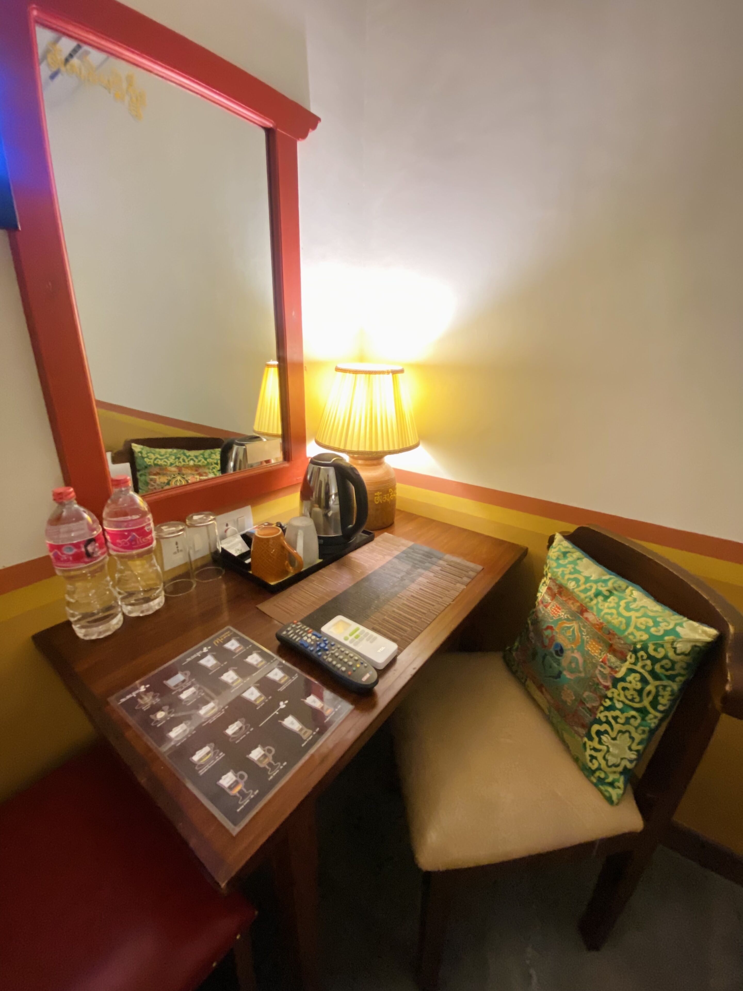 Une chambre d’hôtel avec un bureau en bois, un miroir, une lampe, une télécommande, une bouilloire et un oreiller.