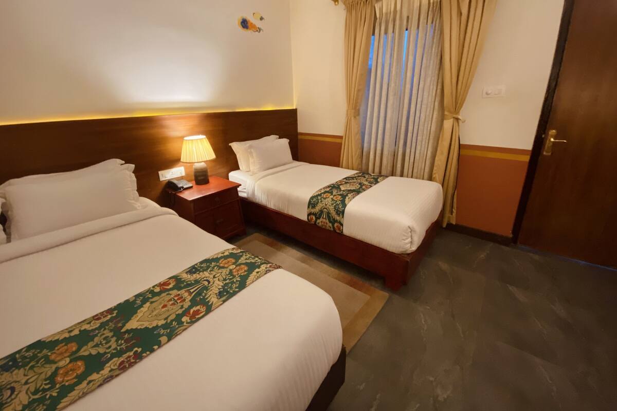 A hotel room with two beds, a nightstand with a lamp, a door, and a window with curtains.