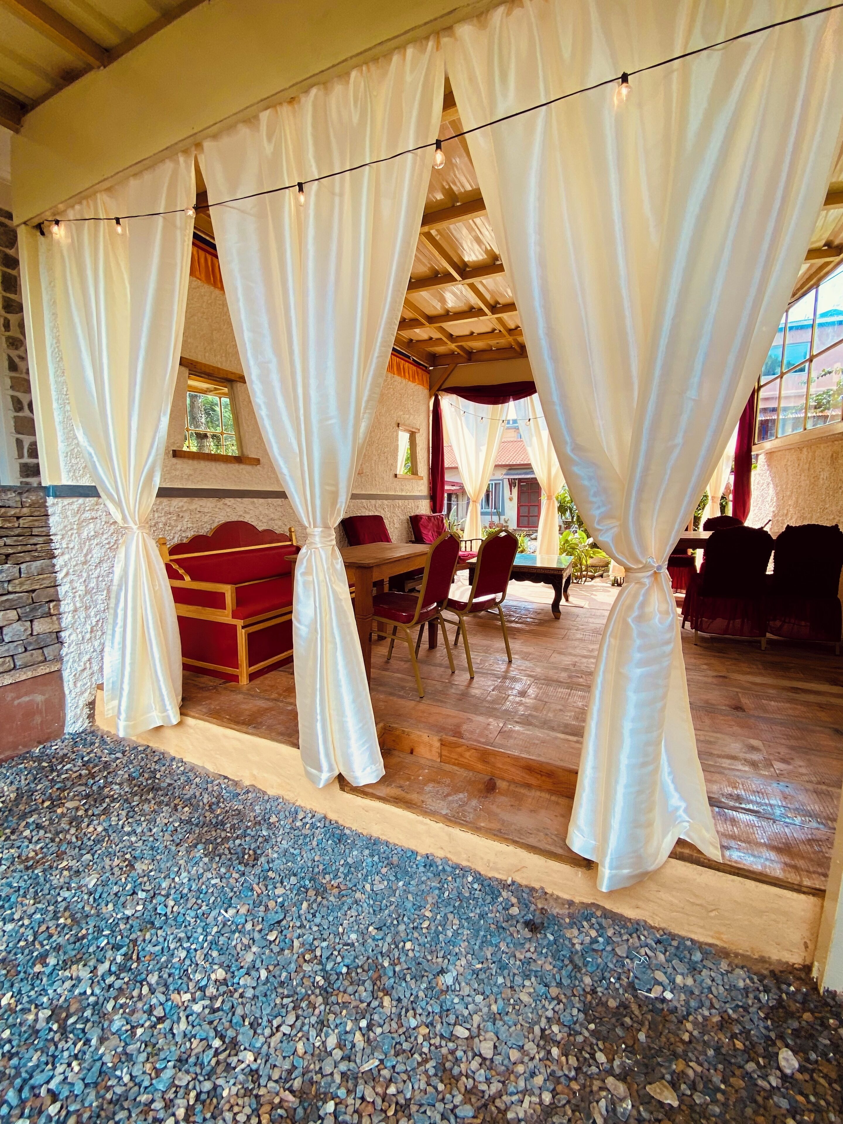 A covered outdoor seating area with a wooden table and chairs, a red sofa, and white curtains.