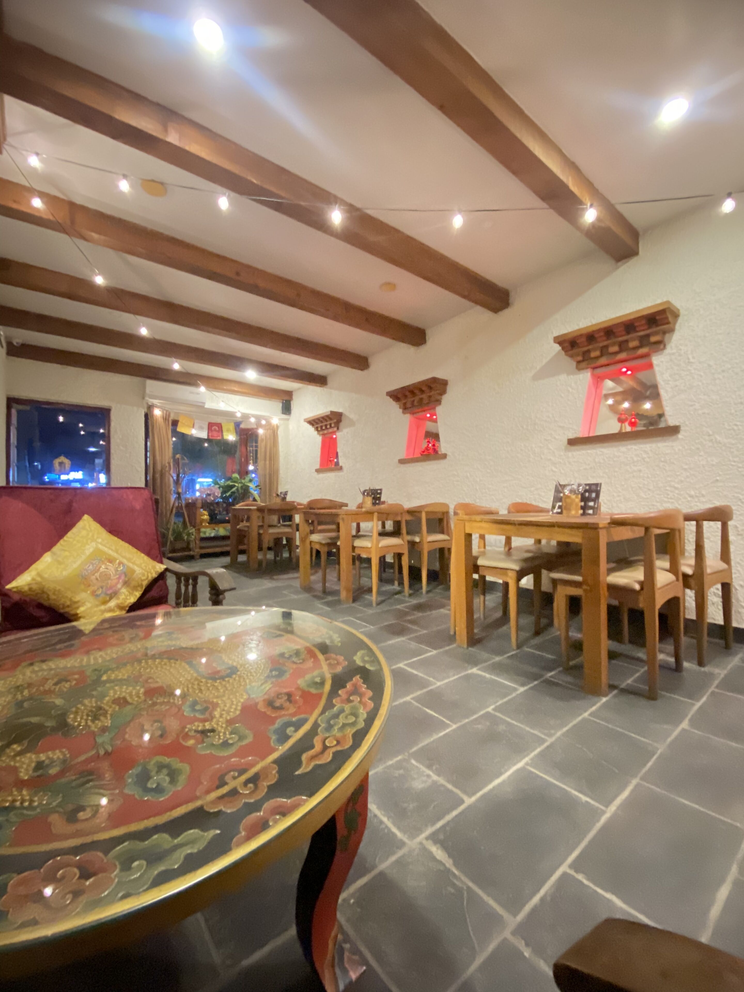 Un comedor con mesas y sillas de madera, una mesa redonda decorativa con motivos florales y un suelo de piedra.
