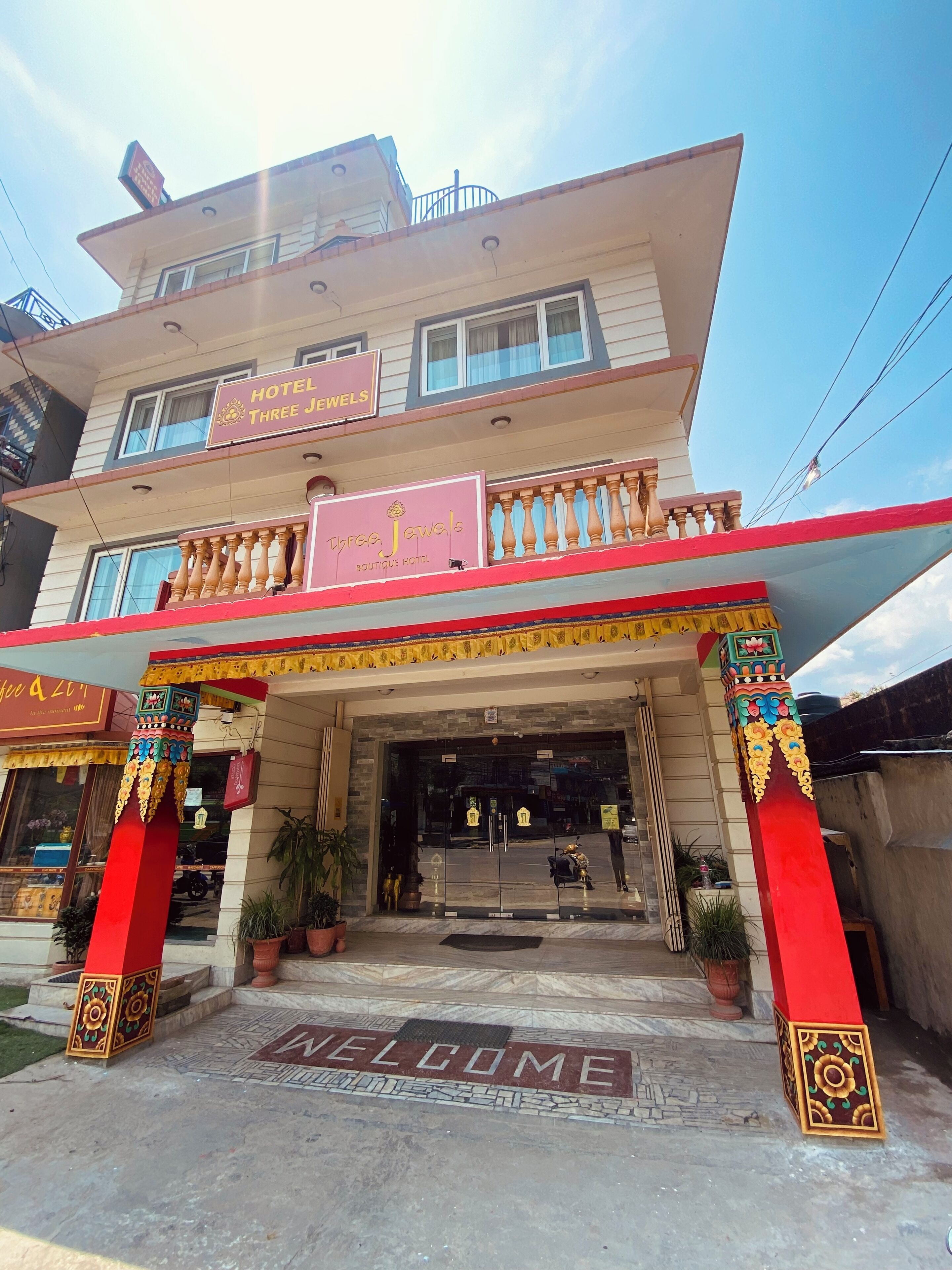 Un hôtel avec une pancarte « Bienvenue » à l’entrée, un balcon doté de balustrades et une enseigne indiquant « Three Jewels Boutique Hotel ».