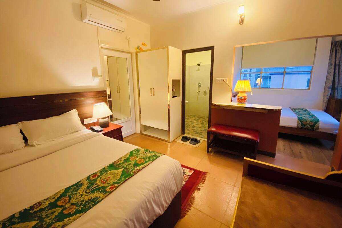 A hotel room with a bed, a desk, a bench, a mirror, a shower, and a window.