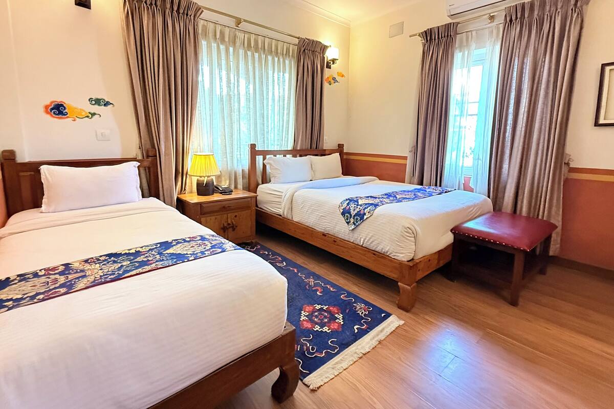 A hotel room with two beds, a nightstand with a lamp, a bench, and a window with curtains.