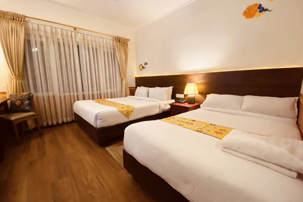 A hotel room with two beds, a desk, a chair, and a nightstand with a lamp.