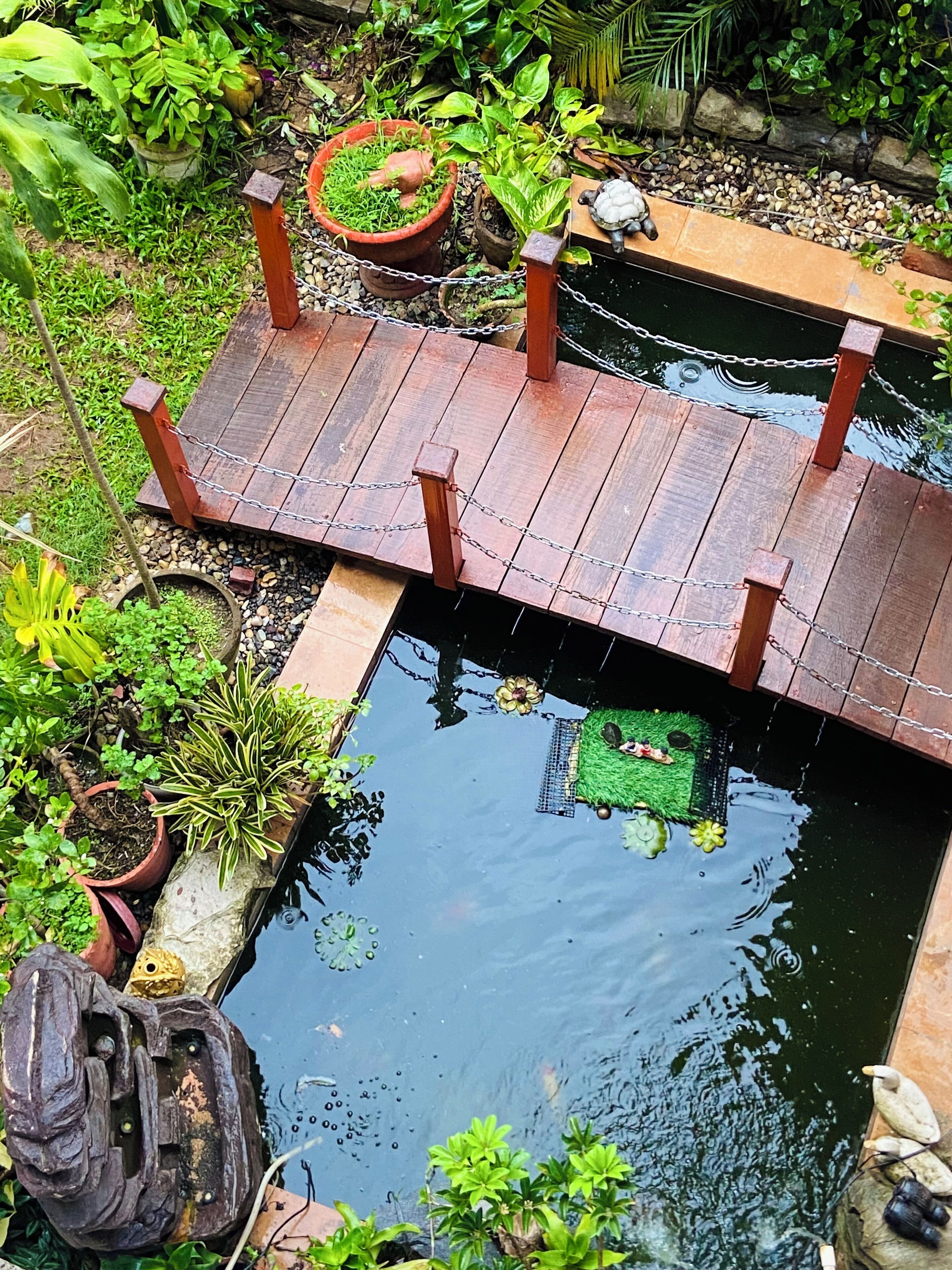 A wooden bridge over a pond with surrounding greenery and potted plants.