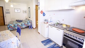Cocina compacta con estufa, lavamanos y refrigerador. Se ve una cama con una colcha colorida al fondo.