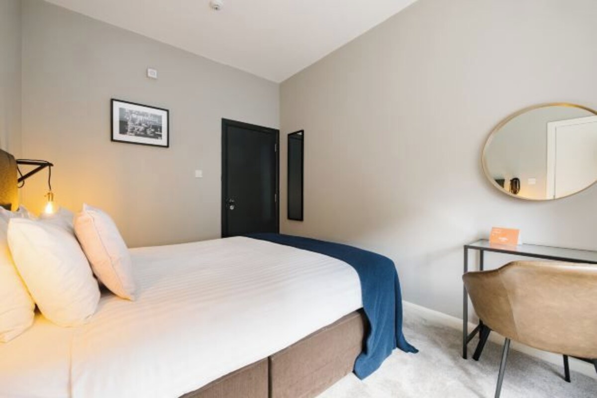 A bedroom with a bed, bedside table, chair, and a mirror on the wall.