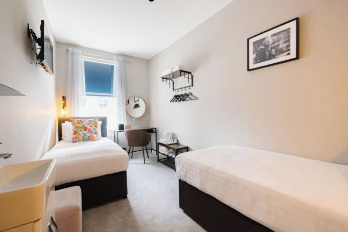 A hotel room with two beds, a desk, a chair, a wardrobe, a mirror, and a window with curtains.
