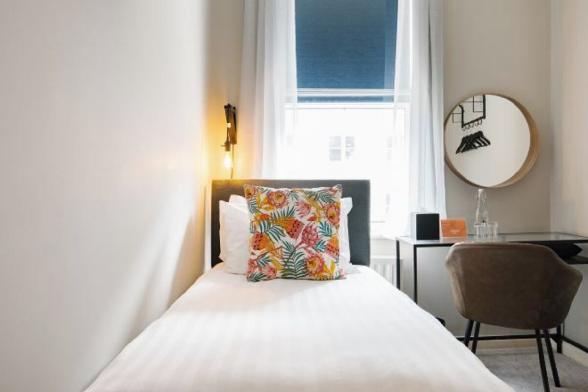 Ett hotellrum med en säng, ett skrivbord, en stol, en rund spegel och ett fönster med gardiner.