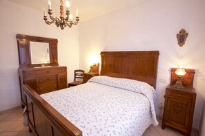 A bedroom with a wooden bed, a nightstand with a lamp, a dresser with drawers, a chair, and a chandelier. - Appartamento La Casina (Portoferraio)
