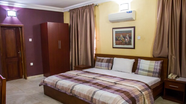 A bedroom with a large bed, a wardrobe, a bedside table with a lamp, and an air conditioning unit on the wall. - Serendipity Anchotel (Lagos)