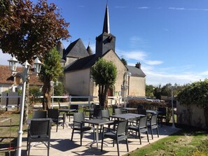 An outdoor seating area with tables and chairs, a church with a pointed steeple, and a clear blue sky. - Le Lanthenay (Romorantin-Lanthenay)