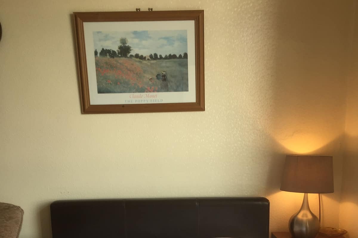 A neatly made bed with white linens, a framed painting of a field above it, a bedside lamp, and a small wooden nightstand with drawers.