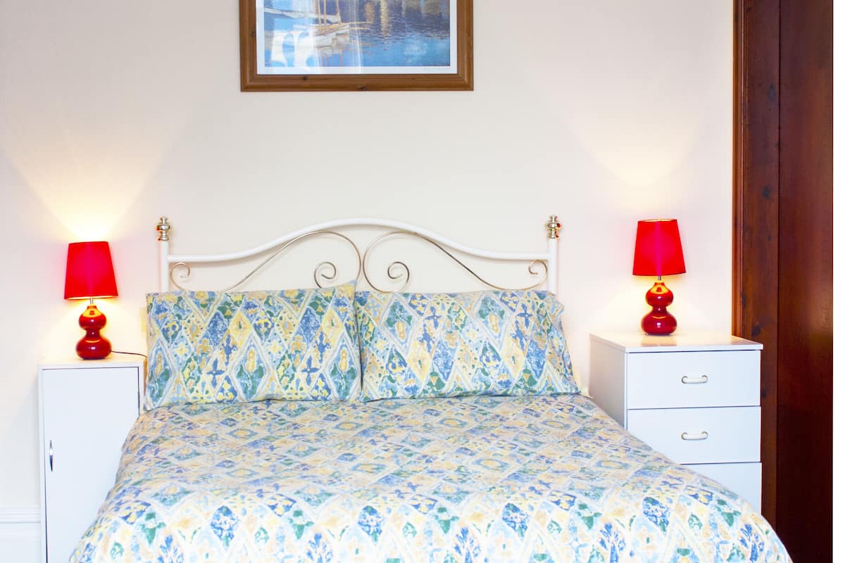 A bed with a patterned bedspread, two bedside tables with lamps, a white headboard, and a framed picture on the wall.